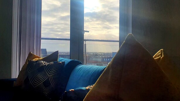 A room with a view through large windows, featuring a blue sofa with patterned cushions and a yellow throw pillow.