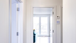 A hallway with white walls and wooden flooring, leading to a room with a large window and a ceiling light fixture.