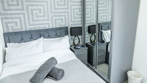 A neatly made bed with white linens, a grey headboard, and a grey blanket. There is a small bedside table with a lamp and a mirror.