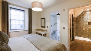 Une chambre à coucher comprenant un lit, une table de chevet, une fenêtre avec des rideaux, un luminaire suspendu, ainsi qu’une salle de bain avec des carreaux décoratifs et une baignoire.