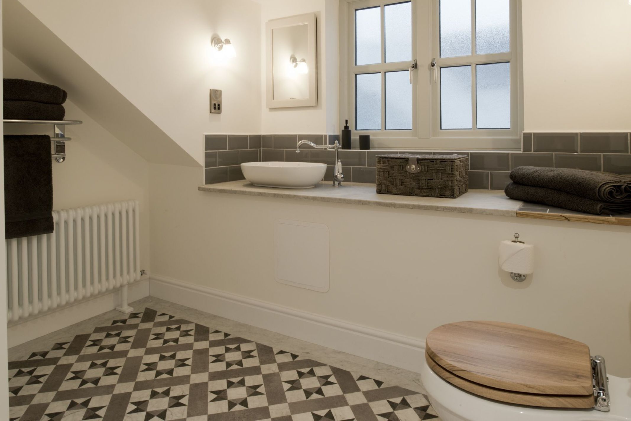 Et badeværelse med en skålhåndvask, toilet, håndklædestang, et vindue og et mønstret gulv.