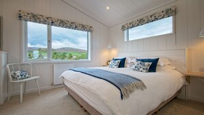 A bedroom with a large bed, two windows with floral curtains, a white chair, and a view of green hills.
