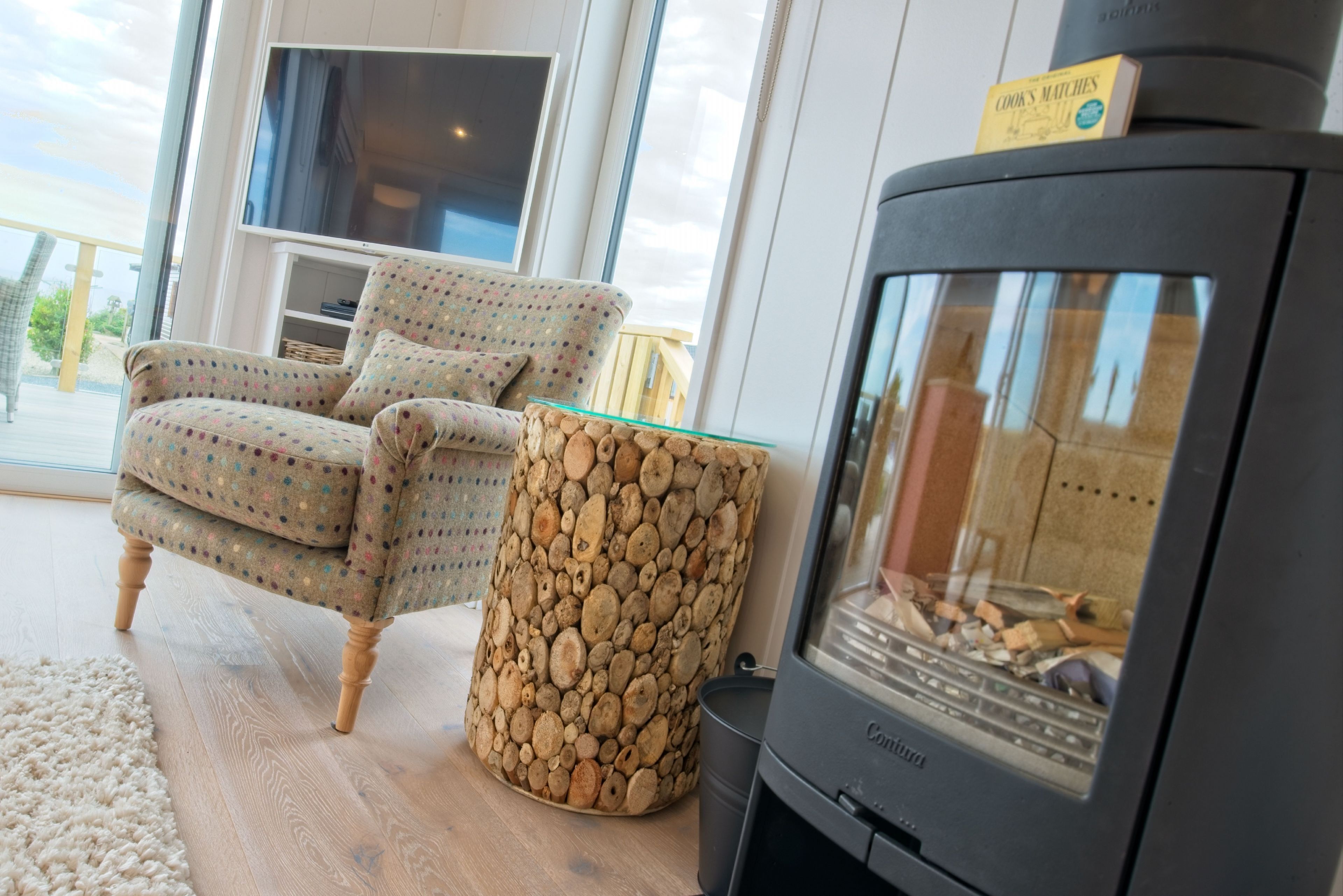 A cozy living room with a patterned armchair, a glass coffee table, and a wood-stone side table. A black fireplace with a book titled "COOK'S MATCHES" is visible.