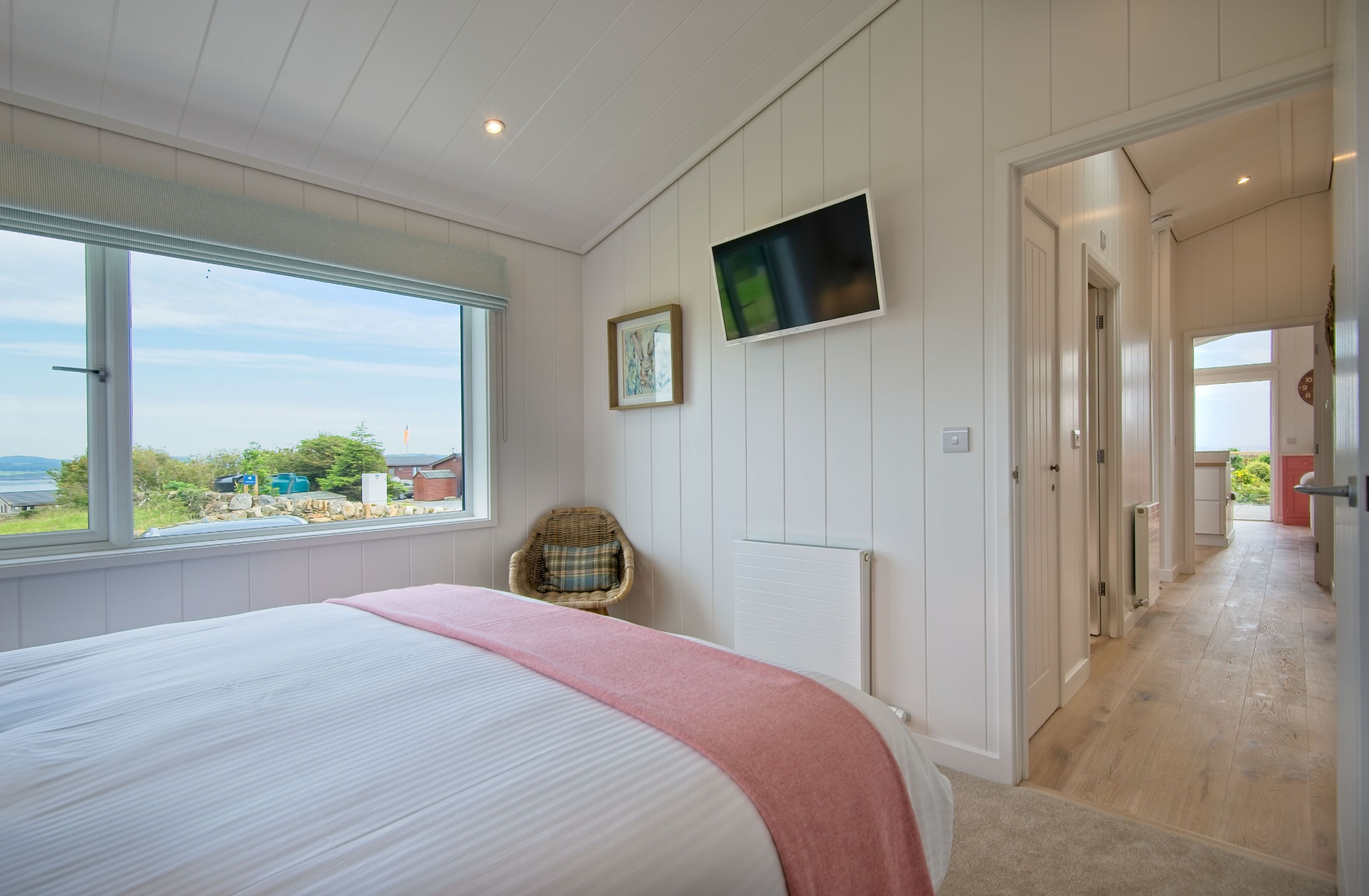 A bedroom with a large bed, a television mounted on the wall, a wicker chair, and a view of the outdoors.