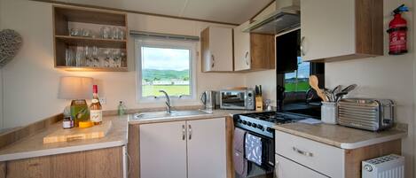 A compact kitchen with a stove, microwave, and sink, wooden cabinets, and a window offering a view of the outdoors.