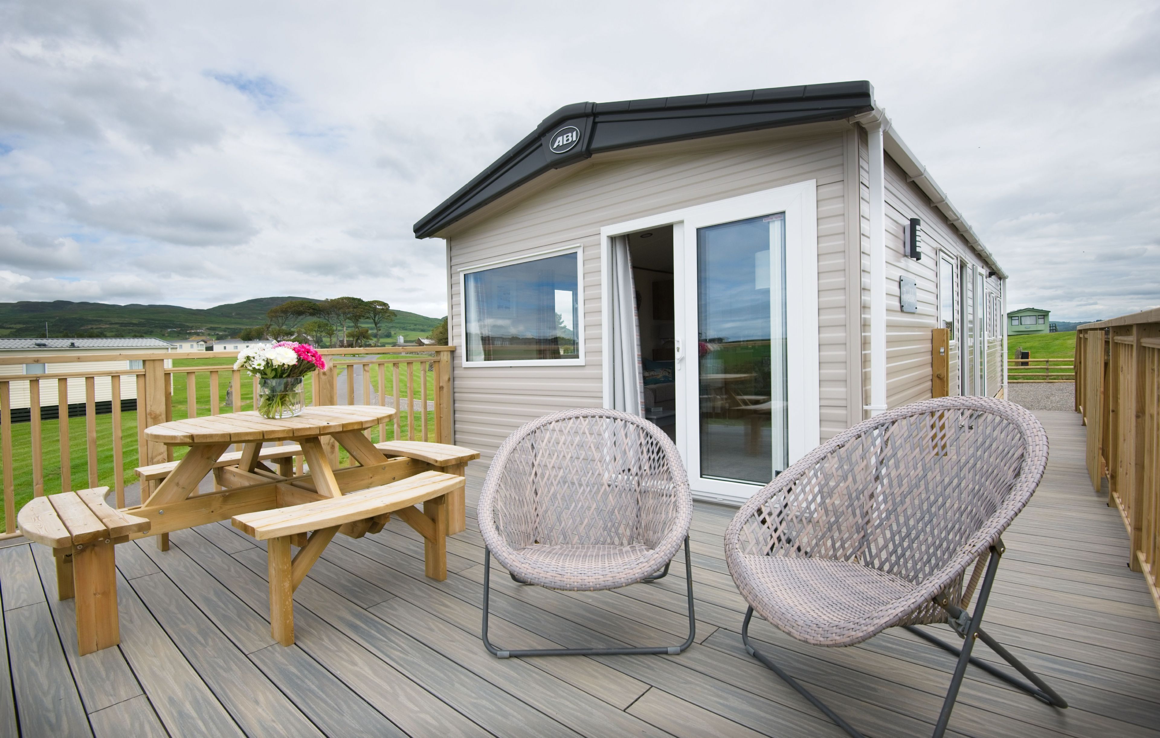 A wooden deck with a picnic table and wicker chairs, a small building with a sliding door, and a view of green hills in the background.