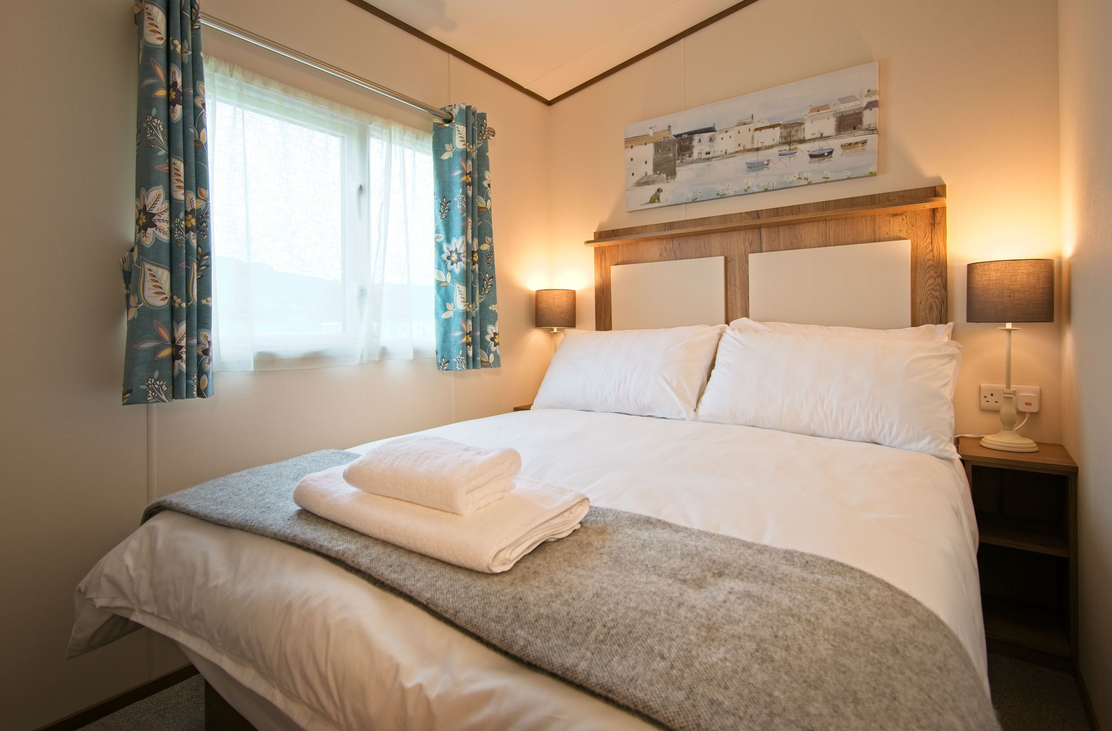 A neatly made bed with white linens and a folded towel, a bedside lamp, and a painting above the headboard.