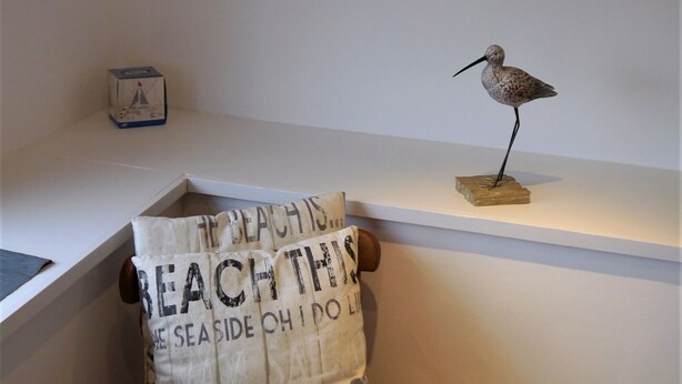 Un coin de pièce avec un fauteuil blanc, une horloge murale décorative et une petite sculpture d’oiseau posée sur une étagère.
