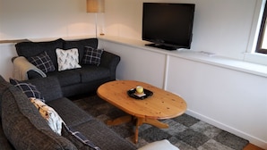Un salon moderne avec un canapé d’angle gris, une table basse en bois, un téléviseur à écran plat fixé au mur et une fenêtre laissant entrer la lumière naturelle.