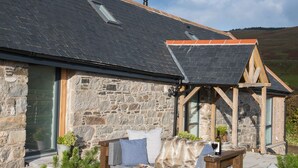 A stone building with a sloped roof, a wooden porch, and a seating area with cushions and a bench. - Dairy Cottage -  a cottage that sleeps 4 guests  in 2 bedrooms (Gatehouse of Fleet)