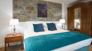 A bedroom with a stone wall, a bed with teal bedding, two bedside tables with lamps, and a wooden wardrobe. - Dairy Cottage -  a cottage that sleeps 4 guests  in 2 bedrooms (Gatehouse of Fleet)