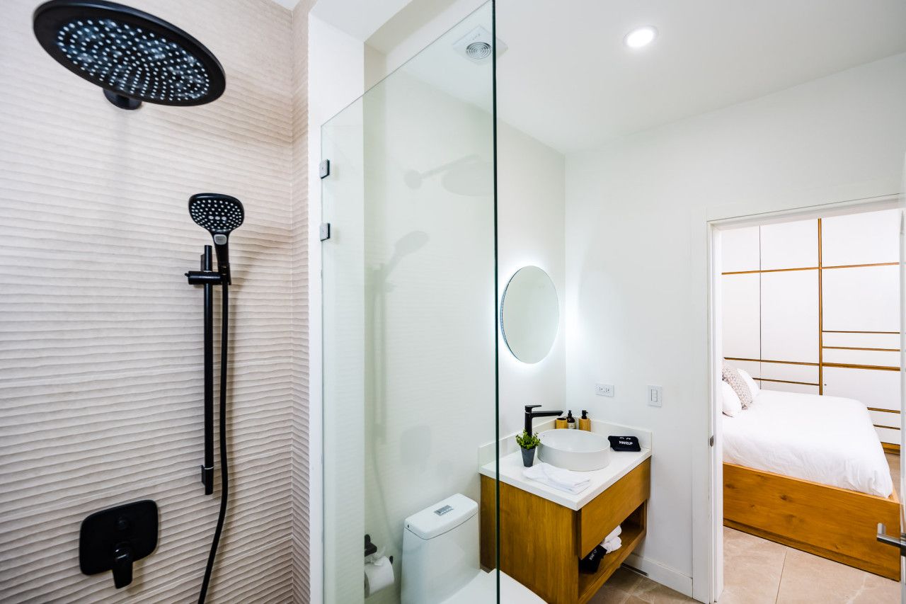 Un baño moderno con cabina de ducha de vidrio, un lavabo blanco con espejo y una cama de madera en la habitación contigua.