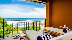 A balcony with two lounge chairs, a small table, and a magazine. The balcony overlooks a beach with greenery and the ocean.