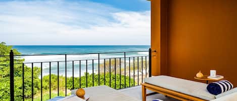A balcony with two lounge chairs, a small table, and a magazine. The balcony overlooks a beach with greenery and the ocean.