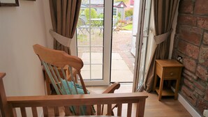 Una habitación con una cama de madera, una silla de madera, una mesita de noche pequeña de madera y un ventanal corredero que da a un área exterior con un patio de ladrillo y una mesa con sillas.