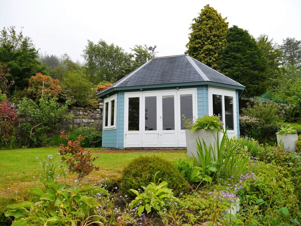 Ein kleines, hellblaues und weißes Wintergartenhaus mit spitzer Dachform, umgeben von üppiger Gartenbepflanzung.