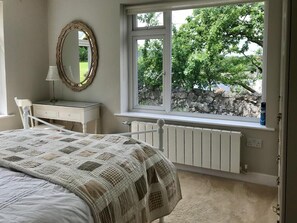 Une chambre à coucher comprenant un lit, une commode, un miroir et une fenêtre donnant sur des arbres et un mur de pierre.