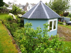 Ein kleines blaues Cottage mit spitzem Dach, umgeben von üppiger Vegetation und einer Steinmauer.
