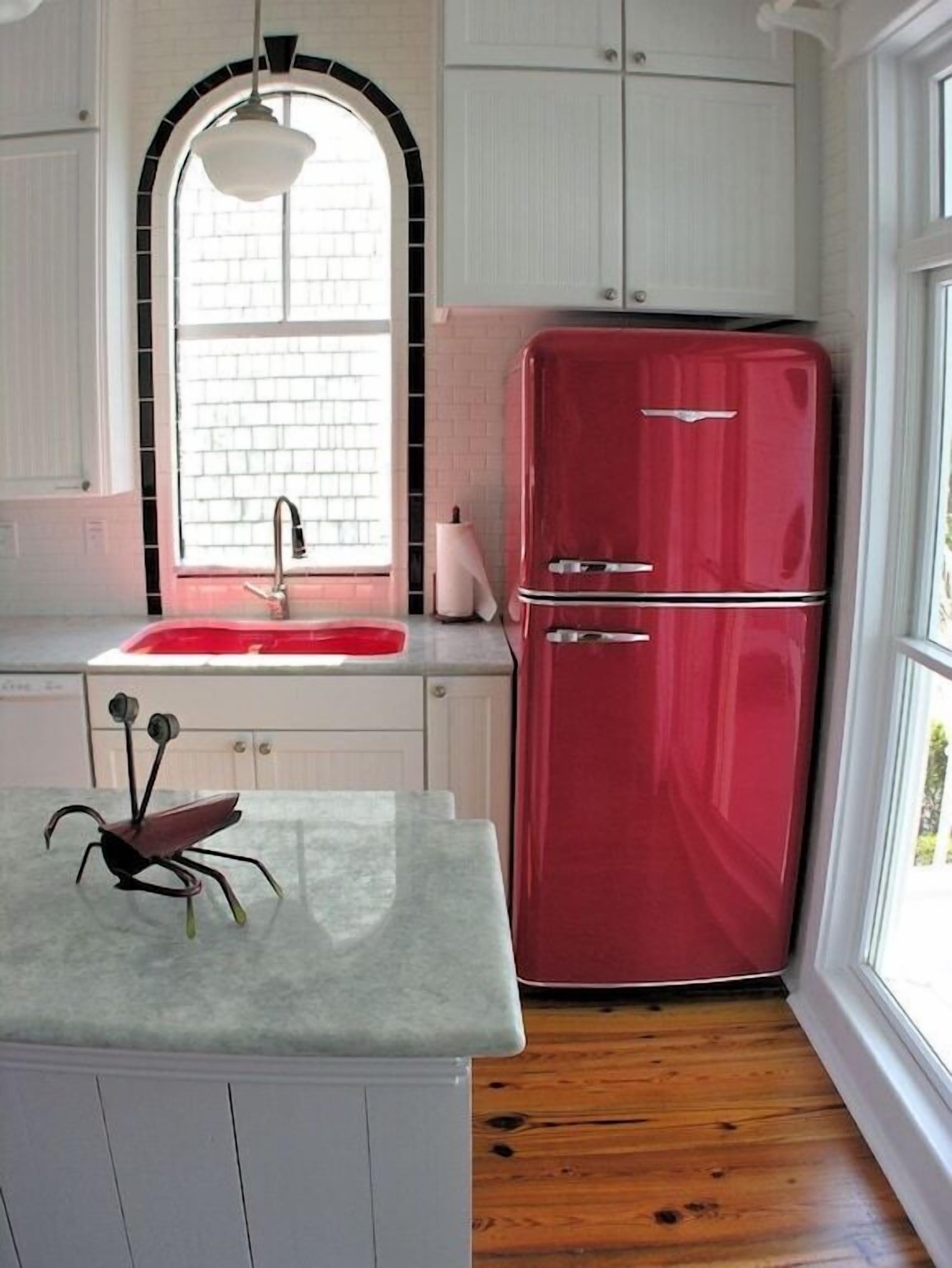 Una cocina con armarios blancos, un refrigerador rojo, una encimera de mármol y una ventana con diseño de ladrillo.