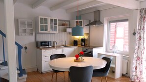 A compact kitchen with white cabinetry, a microwave, a kettle, and a coffee maker. There's a dining table with chairs and a hanging pendant light. - Cozy vacation home with fenced plot, 3 bedrooms, 2 bathrooms (Greetsiel)