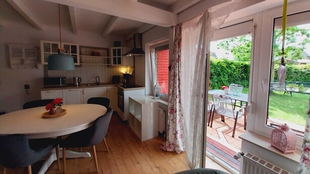 A compact kitchen with modern appliances, a dining area with a round table, and a view of a garden through large windows. - Cozy vacation home with fenced plot, 3 bedrooms, 2 bathrooms (Greetsiel)