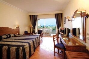 A hotel room with two beds, a desk, a chair, a TV, and a balcony with a view of the beach and palm trees. - Bliss Nada Beach Resort (Marsa Alam)