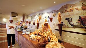 Een buffet met verschillende broden en gebak, koks in witte uniformen, en een muurschildering van kameel.'