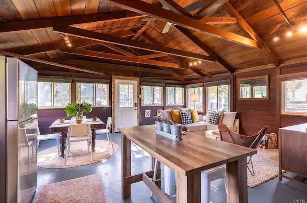 Een ruime kamer met een hoge plafond, houten balken, een zithoek met tafel en stoelen, een zandbank met kussens, en grote ramen voor natuurlijk licht.