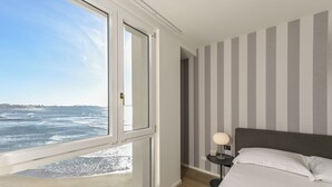 A bedroom with a large window overlooking the ocean, a bed with white linens, a nightstand with a lamp, and striped walls.