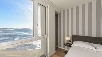 A bedroom with a large window overlooking the ocean, a bed with white linens, a nightstand with a lamp, and striped walls.