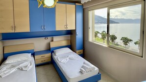A hotel room with a bed, a chest of drawers, a wardrobe, and a window with a view of a lake and mountains.