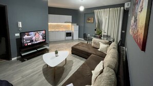 A modern living room with a flat-screen TV, a white coffee table, a brown sofa, and a dining area with a glass table and chairs.
