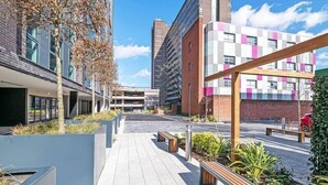 Moderni kaupunkiaukio, jossa on päällystetty kävelyreitti, penkkejä ja istutusastioita.