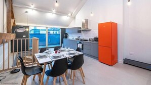 Una cocina moderna con un refrigerador naranja brillante, un área de comedor con una mesa preparada para cuatro comensales y vistas al exterior a través de amplias ventanas.