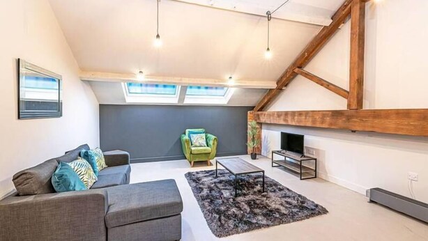 Un salón estilo loft con techo inclinado, tragaluces, un sofá gris, un sillón verde, una alfombra negra y una chimenea.