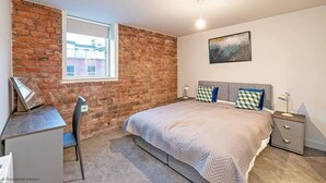 Un dormitorio con pared de ladrillo, cama con almohadas estampadas, escritorio y ventana.