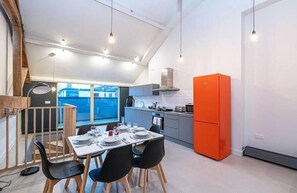 Una cocina moderna con un refrigerador naranja brillante, un área de comedor con una mesa preparada para cuatro comensales y vistas al exterior a través de amplias ventanas.