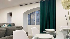 Uma sala de jantar moderna com mesa de vidro e cadeiras brancas, um sofá cinzento e vista para um edifício através de uma janela com cortinas verdes.