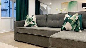 Uma sala de estar moderna com um sofá seccional cinza, almofadas estampadas em verde e branco, uma janela grande com cortinas e um quadro na parede.