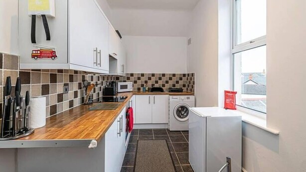 Een kleine keuken met een houten aanrecht, witte kastjes, een magnetron, een wasmachine en een raam.