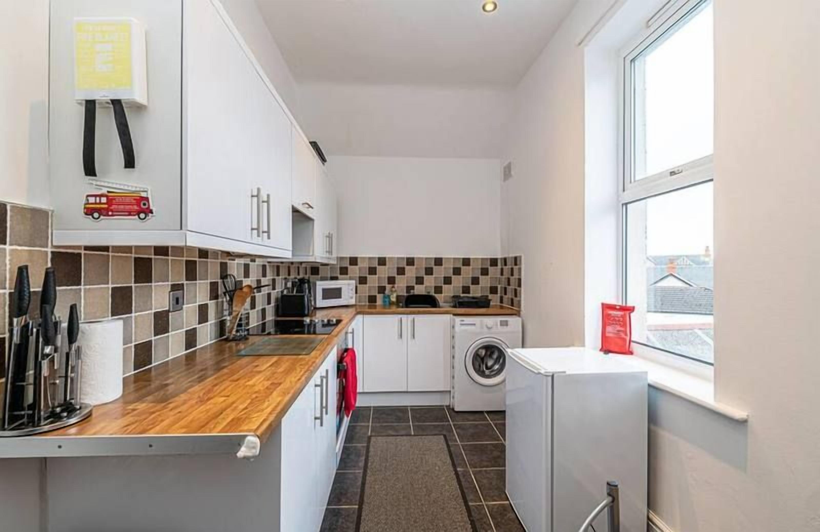 Een kleine keuken met een houten aanrecht, witte kastjes, een magnetron, een wasmachine en een raam.