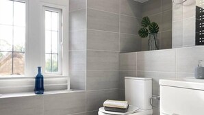 Une salle de bain moderne avec des carreaux gris, des toilettes blanches, une fenêtre ornée d’un vase bleu et un lavabo avec un distributeur de savon.