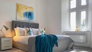 Une chambre à coucher moderne, comprenant un lit, des tables de chevet, une commode, une fenêtre avec des stores et une œuvre d’art accrochée au mur.