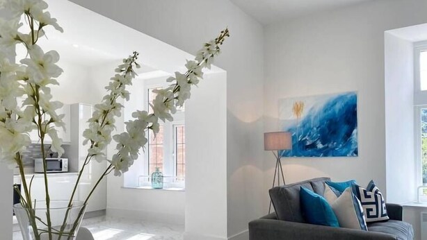 Un salon moderne avec un canapé gris, une table basse en verre et des orchidées blanches. Un tableau est accroché au mur et une lampe se trouve à côté du canapé.