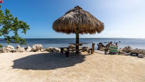 A beach with a thatched umbrella, picnic tables, and a signpost with directions. - Amazing Key Largo Tropical Paradise 3/3 Townhouse with Water Lagoon view (Key Largo)