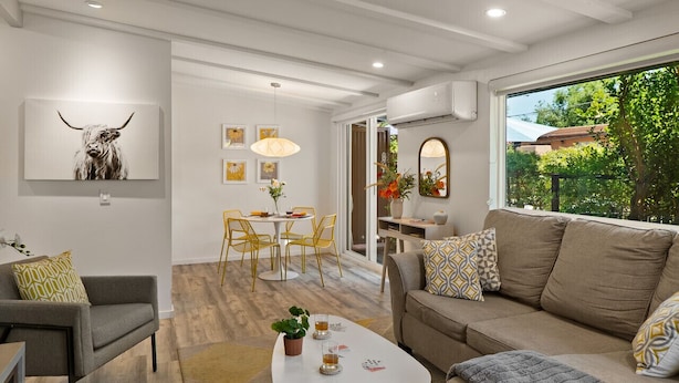 A modern living room with a sofa, armchair, and coffee table. There is a dining area with a table and chairs, and a large window with a view of greenery. - DreamCatcher-S Uptown! Walk to Shop/Dine/Hike (Sedona)