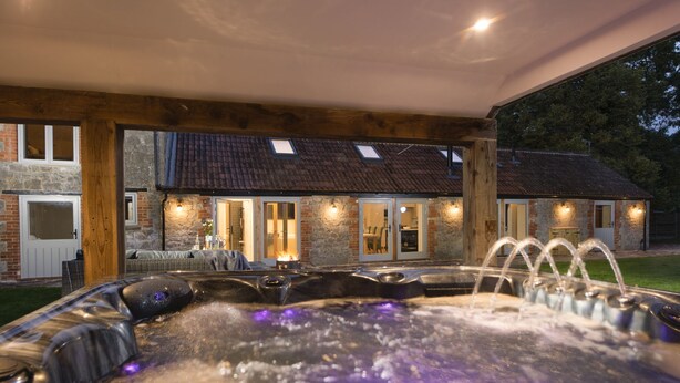 A hot tub area with a stone wall and wooden beams, a covered seating area, and a stone building in the background.