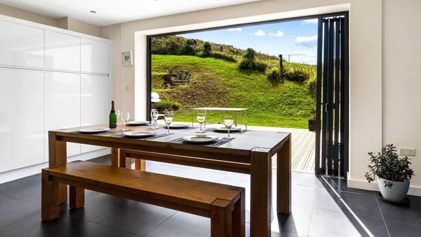Une cuisine moderne avec une grande table à manger pour quatre personnes, un banc et une vue sur un paysage verdoyant par une porte ouverte.