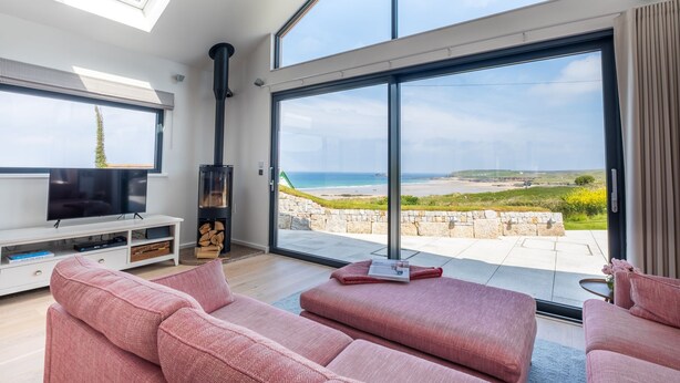 A modern living room with a large TV, pink sofas, a fireplace, and a view of the sea. - Gwithian Lookout -  a house that sleeps 5 guests  in 3 bedrooms (Gwithian)