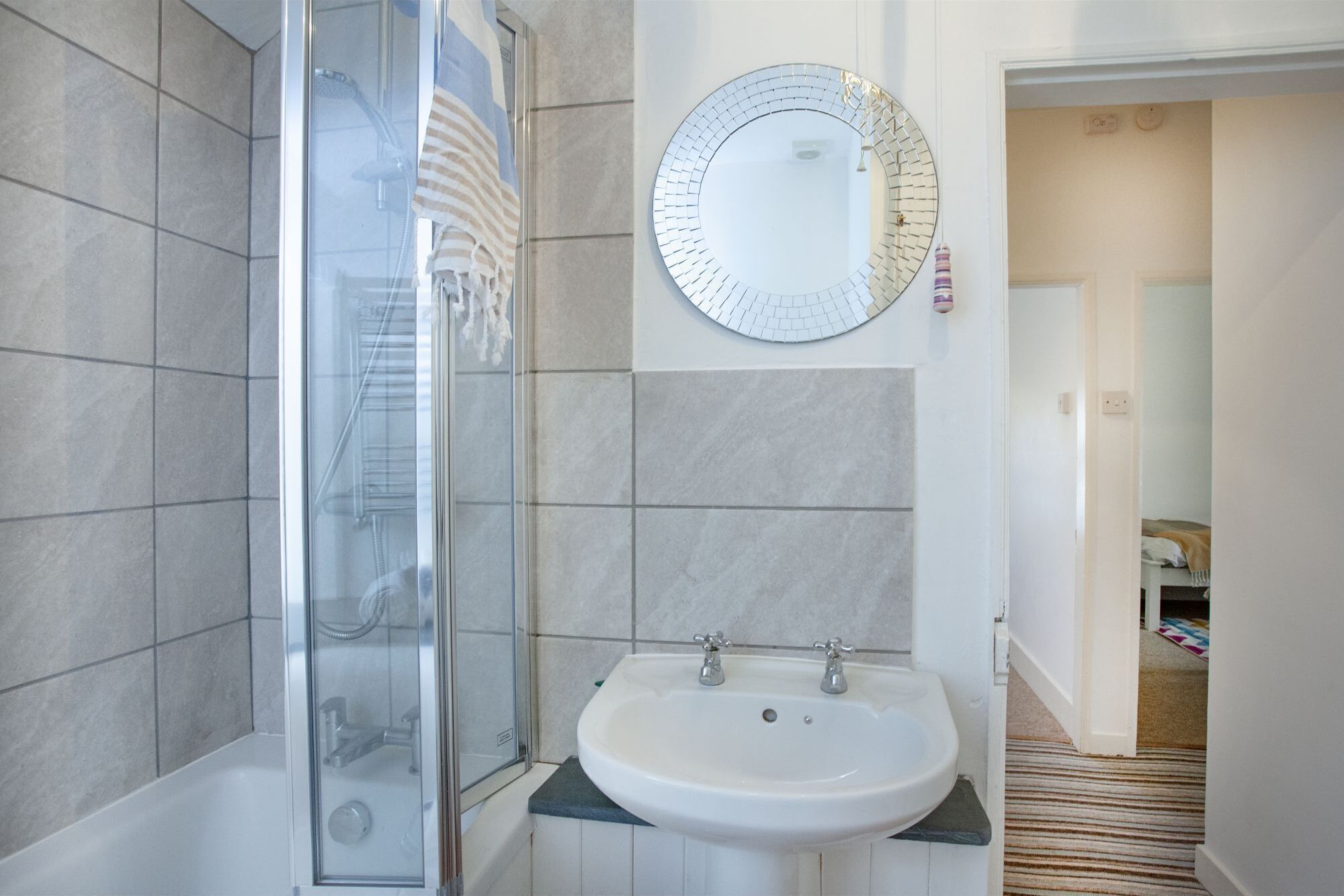 A bathroom with a glass shower enclosure, a white sink with two faucets, and a round mirror with a grid pattern.